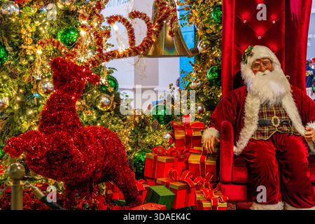 Die Figur des Weihnachtsmanns sitzt auf einem roten Samthron, umgeben von gewickelten Geschenken und einem hell erleuchteten Weihnachtsbaum. Festliche Feiertage wecken Freude und Freude Stockfoto