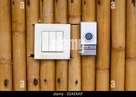 Nahaufnahme eines weißen elektrischen 3-fach-Lichtschalters und einer modernen Türklingel, die nebeneinander auf einer rustikalen, gewebten Bambuswand installiert sind. Kontrast von Stockfoto