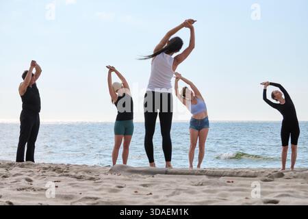 Ein Yogalehrer für Erwachsene führt verschiedene Erwachsene in Dehnungsübungen an der Sandküste der Ostsee unter klarem Himmel, um Wellness und Achtsamkeit zu fördern Stockfoto