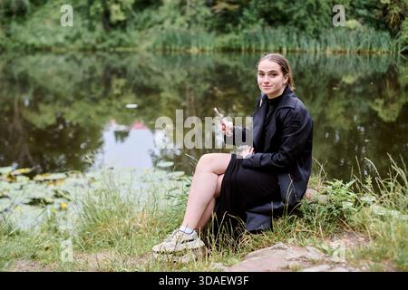 Erwachsene Frau mit braunen Haaren in schwarzem Outfit sitzt auf Gras von Reflektierendem Teich umgeben von Grün. Tageslicht sorgt für eine ruhige, besinnliche Atmosphäre Stockfoto