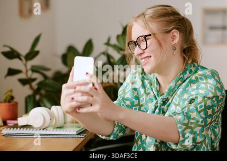 Junge Frau lächelt, während sie Selfie mit Smartphone in Innenräumen nimmt Stockfoto