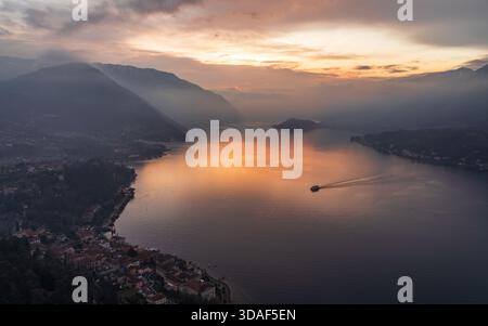 Aus der Vogelperspektive auf goldenes Sonnenlicht, das das ruhige Wasser des Comer Sees küsst, mit Varennas bezaubernder Architektur entlang der Küste, Varenna, Lo Stockfoto
