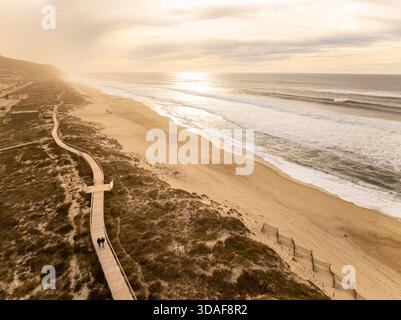 Aus der Vogelperspektive des goldenen Sandes treffen Sie auf die schäumenden Wellen, ein Holzpfad schlängelt sich durch Dünen unter einem weichen, trüben Himmel, Praia de Quiaios, Figueira da Foz, Portuga Stockfoto