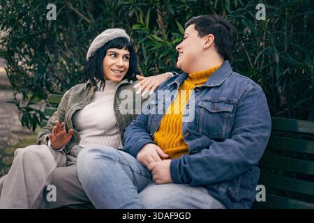 Zwei junge, multiethnische Frauen sitzen auf einer Parkbank und reden, lachen und entspannen sich zusammen. Stockfoto