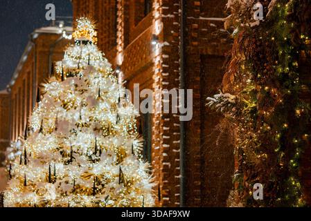 Der hell beleuchtete weihnachtsbaum mit warmweißen Lichtern und Schnee steht vor einem Backsteingebäude und schafft eine festliche Winterszene an einem schneebedeckten Feiertagsabend Stockfoto