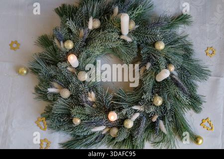 Ein schneebedeckter Weihnachtskranz, verziert mit goldenen Kugeln und weißen Kerzen, mit zwei brennenden Kerzen. Stockfoto