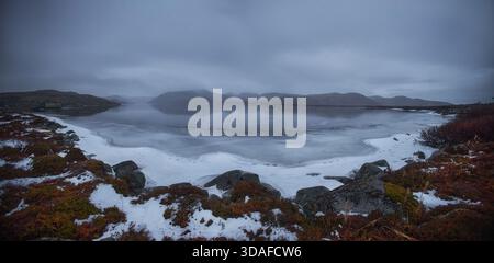 Eine verschneite Szene in Teriberka zeigt einen See, der von Hügeln und Felsen unter einem bewölkten Himmel umgeben ist. Die Gegend zeigt die Schönheit des Winters in Russland. Stockfoto