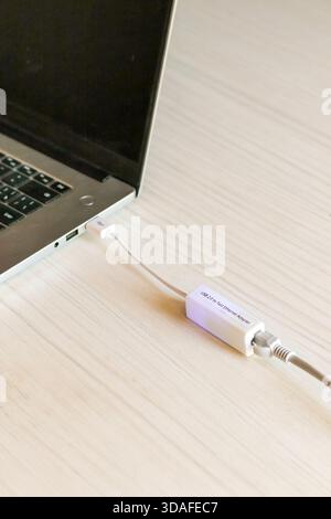Nahaufnahme eines silbernen Laptops, der mit einem weißen USB-Gigabit-Ethernet-Adapter verbunden ist und über ein Netzwerkkabel angeschlossen ist, und auf einem hellen Holzpult für Re Stockfoto