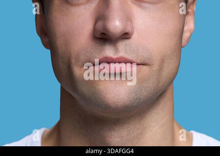 Kaukasischer junger erwachsener Mann, der die untere Gesichtshälfte mit neutralem Ausdruck vor blauem Hintergrund zeigt, Lippen geschlossen, Stoppeln sichtbar, lässiges Hemd trägt Stockfoto