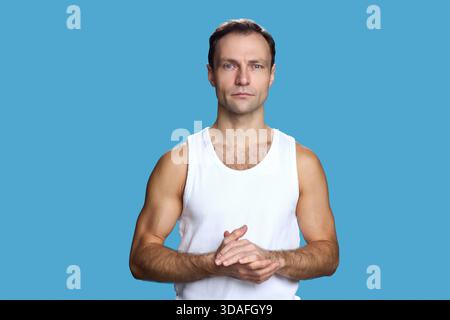 Porträt eines kaukasischen Mannes mittleren Alters, der vor blauem Hintergrund steht, die Hände zusammenfasst und mit neutralem Gesichtsausdruck direkt in die Kamera blickt Stockfoto