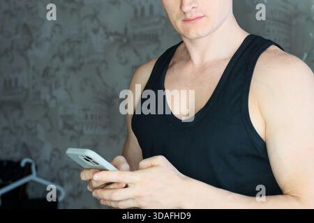 Robustes, männliches Smartphone mit schwarzem Tanktop, das in der häuslichen Umgebung auf das Display starrt Stockfoto