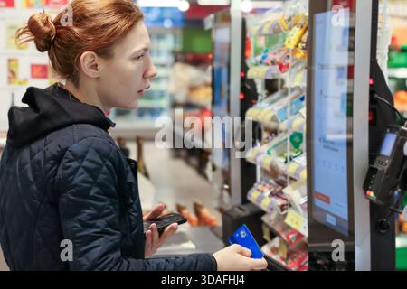 Rothaarige Frau bereitet sich vor, mit Karte zu bezahlen, während sie ihr Smartphone in der Nähe des Imbissregals benutzt Stockfoto