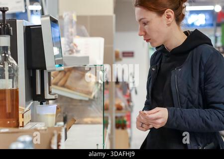 Rothaarige Frau in Jacke, die neben dem Getränkespender mit Tasse im Supermarkt steht Stockfoto