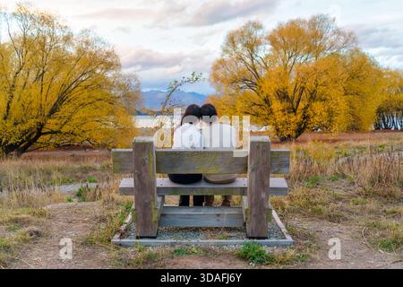 Romantisches Paar in weißem Pullover auf Holzbank, umgeben von goldenen Herbstbäumen mit Blick auf den Lake Tekapo und die Berge in Neuseeland Stockfoto