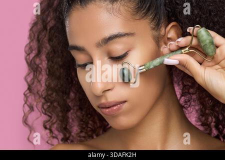 Young adult Black woman using jade roller on cheek, showing skincare routine and facial massage technique, closeup of face with natural makeup, curly hair framing face Stockfoto