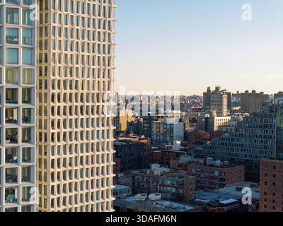 Die scharf weiße Fassade eines modernen Hochhauses aus der Vogelperspektive steht im Kontrast zum warmen Leuchten der Gebäude in der Ferne, Brooklyn, New York, Unit Stockfoto
