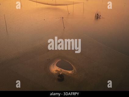 Aus der Vogelperspektive des goldenen Sonnenlichts, das das ruhige Wasser küsst und lange Schatten über die traditionellen Fischernetze und Boote wirft, Hoai Nhon, Binh Dinh, VI Stockfoto