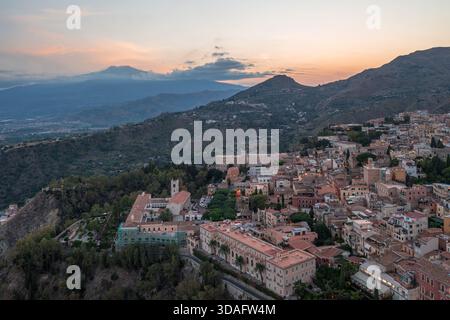Blick aus der Vogelperspektive auf die alten Gebäude, die sich in den Hügel schmiegen, mit dem Ätna, der in der Ferne unter einem lebendigen Sonnenuntergangshimmel ragt, Taormina, Sizilien, Italien Stockfoto