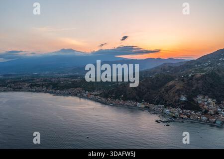 Blick aus der Vogelperspektive auf die Küste und das Meer, mit der Silhouette des Ätna in der Ferne vor einem lebendigen Sonnenuntergangshimmel, Taormina, Sizilien, Italien. Stockfoto