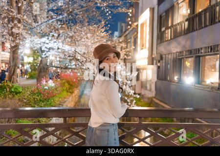 Fröhliche asiatische Frau trägt Hut und genießt auf einer beleuchteten Brücke unter Kirschblüten am Kanal in Gion im Zentrum von Kyoto, Japan Stockfoto