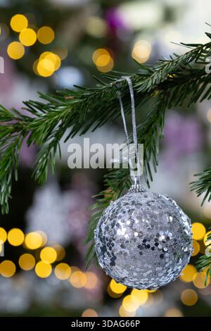 Silberne glitzernde Weihnachtskugel, die an einem Kiefernzweig hängt, mit warmen goldenen Bokeh-Lichtern im Hintergrund, elegante festliche Weihnachtsdekoration Stockfoto