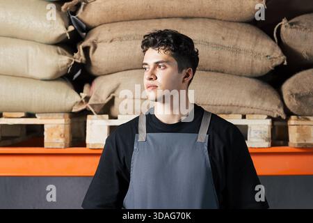 Porträt eines jungen Mannes, der eine Schürze trägt und in einem Lagerhaus vor Säcken mit Kaffeebohnen steht Porträt eines jungen Mannes, der eine Schürze trägt und in A steht Stockfoto