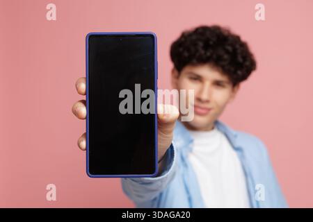 Porträt eines jungen Mannes, der ein modernes Smartphone mit leerem schwarzen Bildschirm auf rosa Hintergrund zeigt, ideal für Mockup von Social Media Online-Diensten für mobile Apps Stockfoto