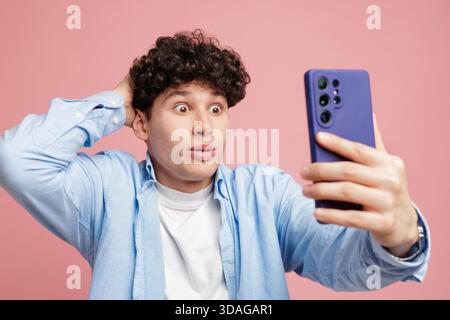 Ein junger Mann mit lockigen Haaren sieht schockiert auf dem Smartphone-Bildschirm aus, was darauf hindeutet, dass Nachrichten, viraler Post oder dringender Anruf auf pastellrosa Hintergrund lokalisiert werden Stockfoto