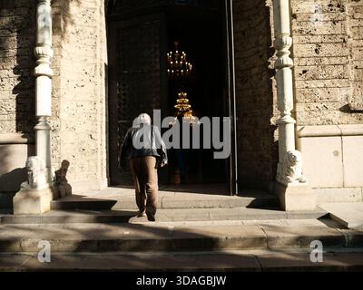 Blick von hinten auf eine ältere Person in einer schwarzen Lederjacke und braunen Hosen, die Steintreppen hinauf in eine historische Steinkirche durch ein großes Stück hinaufgeht Stockfoto
