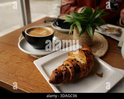 Nahaufnahme mit Schokoladencroissant und zwei Tassen Kaffee mit Latte Art auf Holztisch im gemütlichen Café. Warmes Frühstück am Morgen Stockfoto