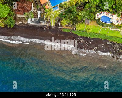 Drohnenblick auf einen vulkanischen Strand mit schwarzem Sand und Luxusresort in Tulamben, Bali Stockfoto