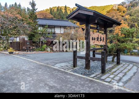 Das Eingangstor zum Ryokan Hanaya (渋本屋) aus dunklem Holz mit geschnitzten Schriften; im Hintergrund das Hauptgebäude mit Sitzgelegenheiten im Freien, umgeben von Bäumen in ihren Herbstfarben, Nagiso, Präfektur Nagano, Japan Stockfoto