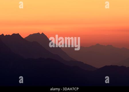 Eine ruhige Berglandschaft bei Sonnenuntergang, mit silbrigen Gipfeln, mehrschichtigen Bergrücken und einem farbenfrohen orangen Himmel, die einen ruhigen und Panoramablick schaffen. Stockfoto