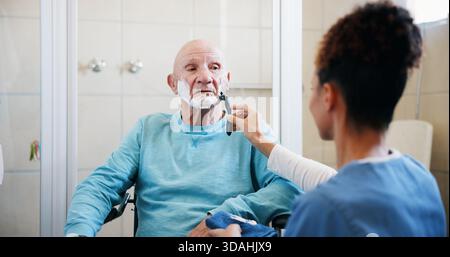 Älterer Mann, Krankenschwester und Rasieren im Badezimmer mit Rollstuhl, Produkt und Schaum für die Pflege zu Hause. Menschen, Betreuungspersonen und Senioren mit Behinderung Stockfoto
