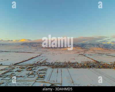 Weite Aussicht aus der Luft auf ein schneebedecktes Dorf und landwirtschaftliche Felder, mit weit entfernten schneebedeckten Bergen, beleuchtet durch goldenes Licht bei Sonnenaufgang oder Sonnenaufgang Stockfoto