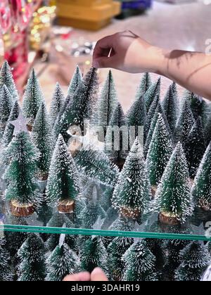 Person arrangiert handwerkliche Dekoration von Miniatur-Weihnachtsbaum-Modellen. Festliche Urlaubsvorbereitung für eine fröhliche Wintersaison mit vielen kleinen, schneebedeckten Stockfoto
