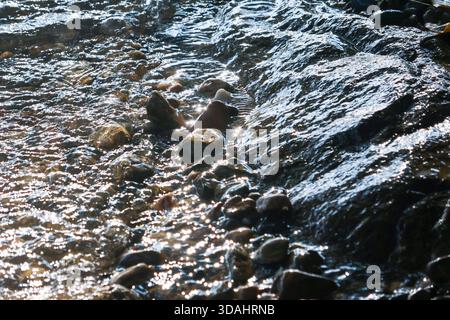 Ruhige Nahaufnahme des klaren Baches, der über glatte Felsen und Kieselsteine fließt. Ruhiges Wasser reflektiert helles Sonnenlicht und schafft einen wunderschönen natürlichen Hintergrund Stockfoto
