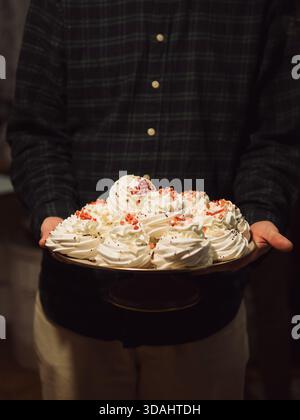 Mann im verwaschenen Hemd hält Meringue-Tablett in warmem, schwachem Licht – vertikale Nahaufnahme Stockfoto