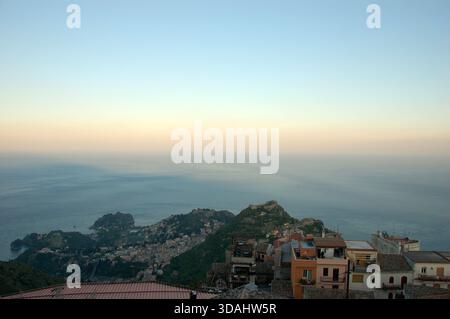 Panoramablick auf die Küste von Taormina, Sizilien, mit Blick auf das Ionische Meer und die Stadt am Hügel bei Sonnenuntergang mit weichem Pastellhimmel und ruhigem Wasser Stockfoto