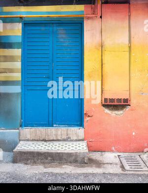 Leuchtend blaue Holztür in einem Gebäude mit gestreiften blauen, gelben, weißen Wänden und orangefarbenen, gelben Abschnitten. Urbane Straßenszene mit gemusterten Stufen, gelber Pfeife und Abfluss. Stockfoto