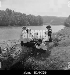 Region Donezk, Ukraine, UdSSR. Mai 1979. Ein authentisches Schwarz-weiß-Archivfoto, das eine Musikstunde im Freien am Ufer zeigt. Ein männlicher Lehrer sitzt auf einem massiven, gefallenen Baumstamm und unterrichtet eine Gruppe von Pionierkindern, die Holzflöten (Sopilkas) spielen. Die Schüler, darunter ein Mädchen in einer Schulschürze und Jungen mit Pionierbande, konzentrieren sich auf ihre Instrumente. Der ruhige Fluss fließt im Hintergrund. Diese Szene fängt die harmonische Mischung aus Natur, Bildung und Kindheit im friedlichen Donbass ein. Stockfoto