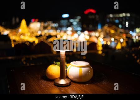 Kleiner, stehender Esstisch für zwei Personen im Freien mit leuchtender Lampe und Obstdekoration in der Nähe eines Weihnachtsmarktes bei Nacht. Nahaufnahme, oberflächlich DE Stockfoto