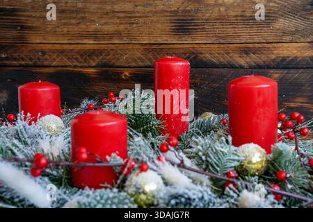 Vier rote Kerzen stehen hintereinander auf einer verschneiten immergrünen Girlande mit roten Beeren und goldenen Kugeln vor rustikalem Holzhintergrund. Stockfoto