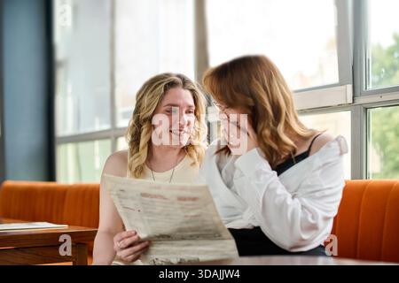 Zwei junge Frauen sitzen dicht auf der orangefarbenen Bank im sonnendurchfluteten Café und teilen sich einen freudigen Moment, während sie sich das Menü ansehen. Ihre entspannte Haltung und ihr Lächeln vermitteln Frien Stockfoto