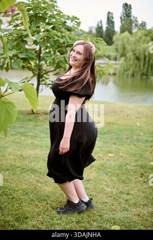 Junge Frau mit braunen Haaren in schwarzem Kleid tanzt glücklich auf dem Grasfeld. Bäume und Teich bilden einen ruhigen Hintergrund. Helles, natürliches Licht verbessert die Stockfoto
