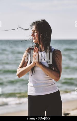 Eine junge Frau mit weißem Tank Top und schwarzer Leggings übt Yoga mit Händen in Gebetsposen am ruhigen Ostseestrand bei Tageslicht, die Augen schlossen mich Stockfoto