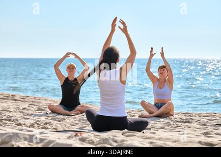 Drei Erwachsene üben sitzende Yoga-Posen auf Strandsand an der Ostsee und Strecken die Arme nach oben. Ruhiges Meer und helles Tageslicht sorgen für eine ruhige, konzentrierte Atmosphäre Stockfoto