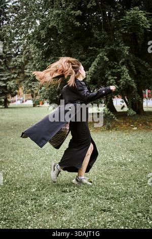 Junge Frau mit blondem Haar dreht sich energisch auf Gras im Park, umgeben von großen grünen Bäumen. Bewegung erfasst mit fließendem Fell und Haar in Natur Stockfoto