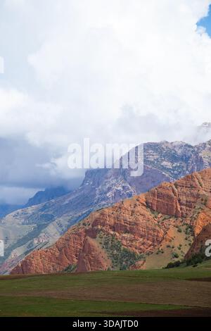 Dramatische Landschaft mit zerklüfteten roten Felsenbergen, die teilweise von Sturmwolken über grünen landwirtschaftlichen Feldern verborgen sind Stockfoto