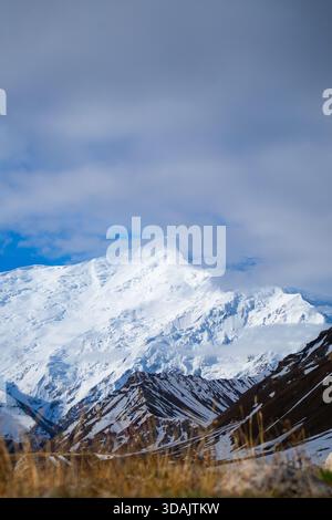 Atemberaubende Aussicht auf einen massiven, schneebedeckten Berggipfel, beleuchtet durch Sonnenlicht unter einem blauen Himmel mit weichen Wolken Stockfoto